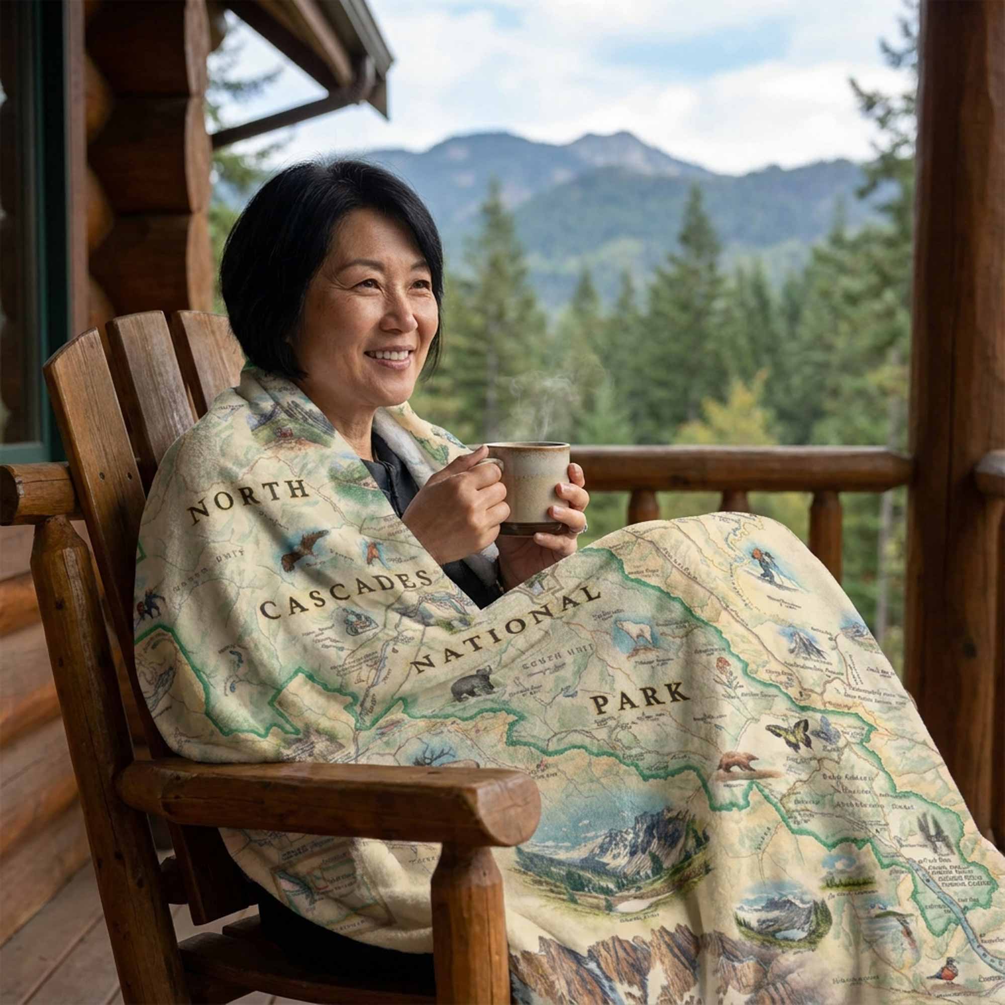 Woman sitting on a wooden chair with a North Cascades National Park blanket, holding a mug, with a scenic background.