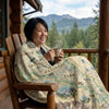 Woman sitting on a wooden chair with a North Cascades National Park blanket, holding a mug, with a scenic background.