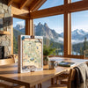 A 1000-piece jigsaw puzzle box titled 'North Cascades' stands on a wooden dining table in a rustic cabin with large windows overlooking a mountain and lake landscape.