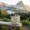 Map-patterned tote bag with a water bottle and guidebook on a wooden bench in a mountainous landscape.