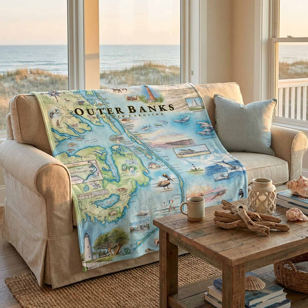 Sofa with a map of Outer Banks, North Carolina blanket draped over it, in a room with large windows overlooking a beach.