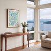 A framed art print of a colorful Port Said map hangs on a white wall in a modern living room with a wooden console table, a vase with greenery, and a large window overlooking a scenic lake and mountains.