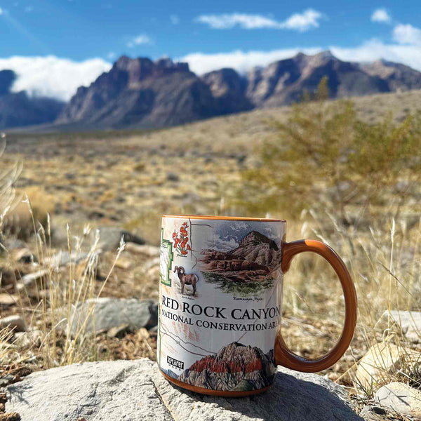 Orange Red Rock Canyon National Conservation Area Ceramic Mug on rock near Red Rocks Canyon. Illustrations: Mt. Willson, Rainbow Mountain, Bridge Mountain, Spring Mountain Ranch. Features red-tail hawk, desert Bighorn, Globemallow, Joshua tree, Prickly pear cactus