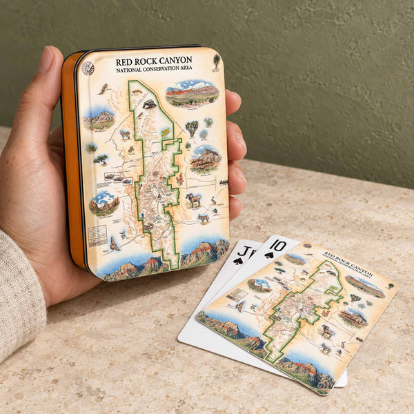 A hand holds a rectangular metal tin and three matching playing cards featuring an illustrated map of Red Rock Canyon National Conservation Area, set on a stone surface against an olive wall.