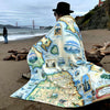 Person sitting on the beach wrapped in a San Francisco map fleece blanket. In the background is the Golden Gate Bridge