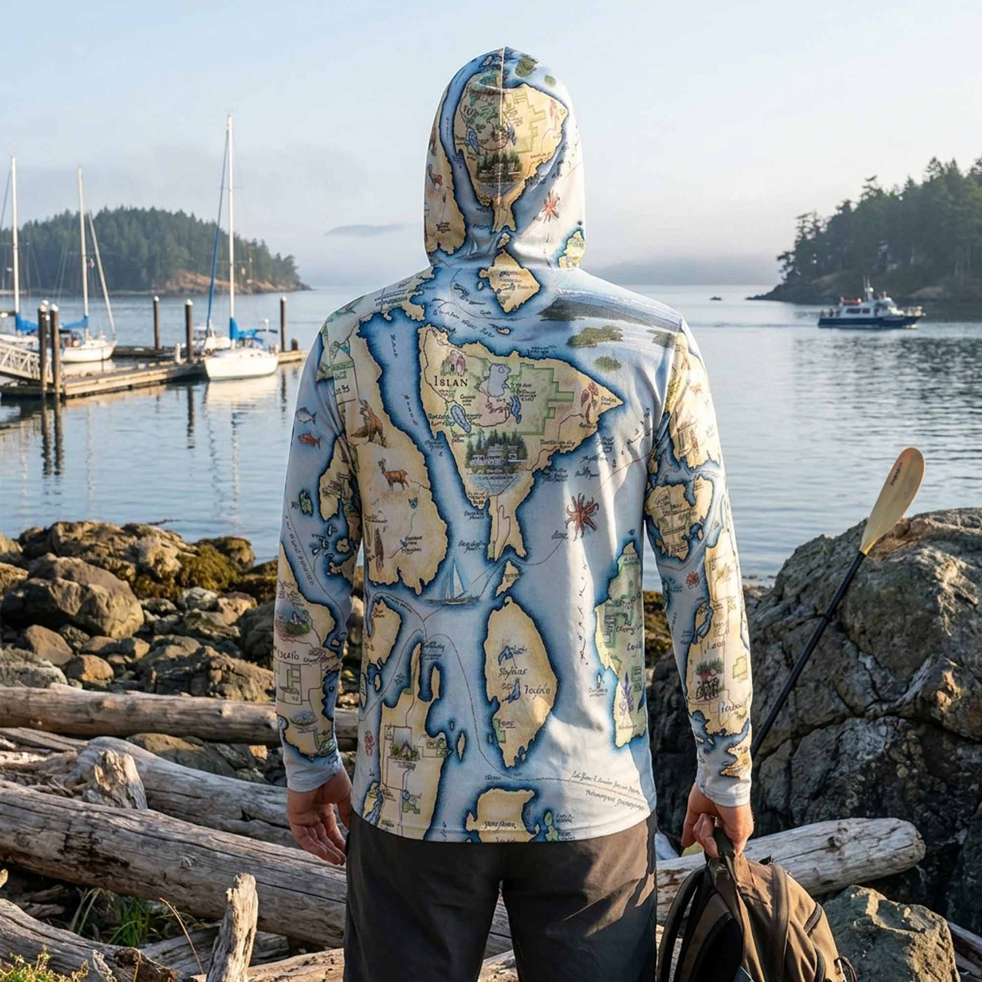 Person wearing a hoodie with a map design by a waterfront with boats and rocks.