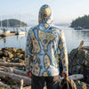 Person wearing a hoodie with a map design by a waterfront with boats and rocks.