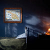 A framed vintage map of Great Gorda Island hanging on a dark blue wall above a bed in a dimly lit bedroom. A bedside table with a lit lamp is visible on the right.