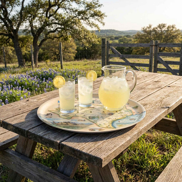 Outdoor setting with a wooden table, pitcher of lemonade, glasses on a Texas servign Tray, and a scenic background.