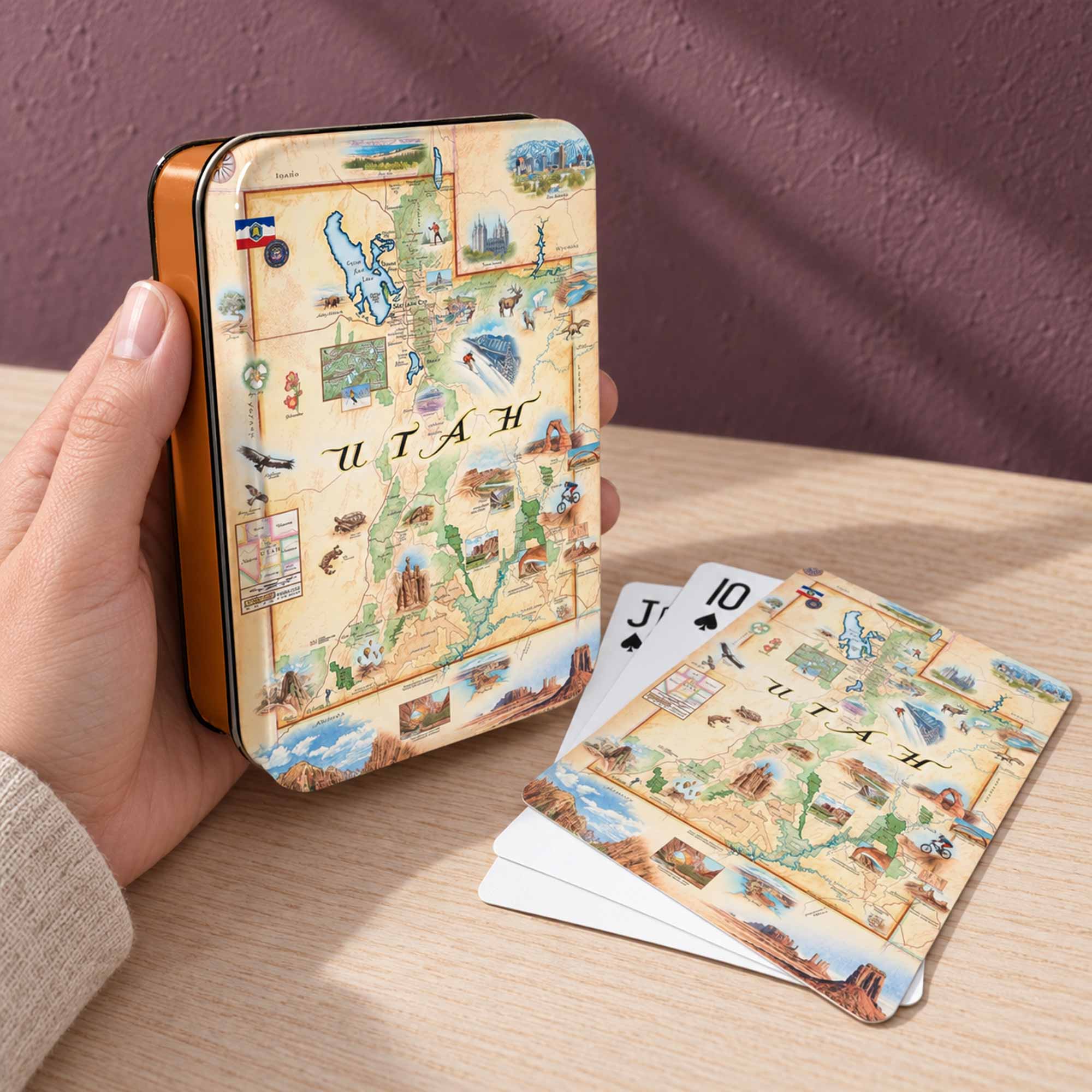 A hand holds a small rectangular tin and several matching playing cards are fanned out on a wooden table. The tin and cards feature a vintage-style illustrated map design labeled Utah, with a warm beige background and multicolored scenic details.