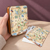 A hand holds a small rectangular tin and several matching playing cards are fanned out on a wooden table. The tin and cards feature a vintage-style illustrated map design labeled Utah, with a warm beige background and multicolored scenic details.