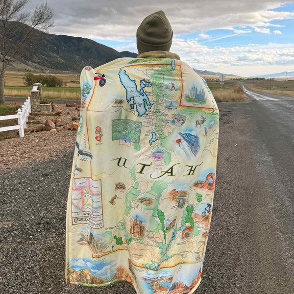 A man wearing Utah State Map fleece blanket around him. Featuring intricate details of popular cities, iconic landmarks such as Arches National Park and Bryce Canyon, and vibrant flora and fauna indigenous to the region.