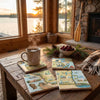 Cup of coffee on a wooden table with scenic coasters, pinecones, and a bowl of fruit in a cozy room with a lake view.