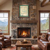 A framed vintage-style map print with a green border hangs above a stone fireplace in a rustic living room with leather sofas, a coffee table, and mountain views through the windows.