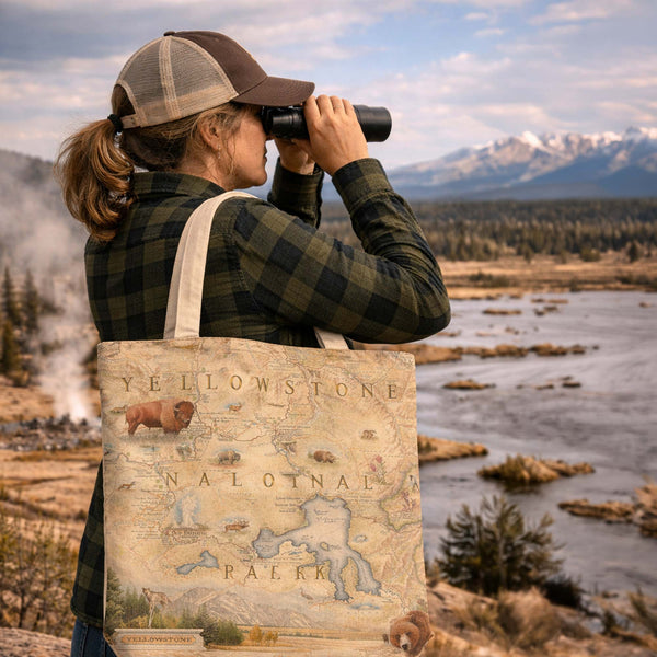 A woman in a brown and beige trucker hat and green plaid shirt looks through binoculars outdoors by a river, carrying a canvas tote bag printed with a vintage map of Yellowstone National Park and an illustration of a bison.