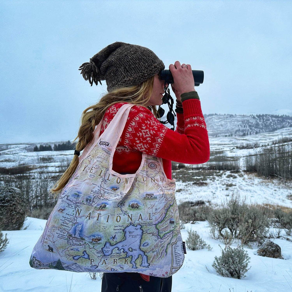 Person in a red sweater looking through binoculars at Yellowstone National Park, carrying a map-themed pouch tote bag, with snow-capped mountains in the background.