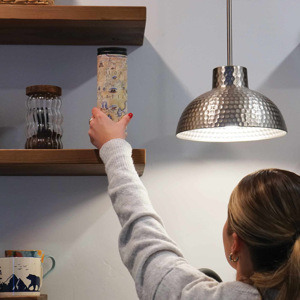 Kitchen scene: Women selecting Yellowstone Travel Mug from wooden shelf. 16 oz mug showcases park icons like Yellowstone Lake, Old Faithful, and Roosevelt Tower, with wildlife illustrations including grizzly bears and mountain lions