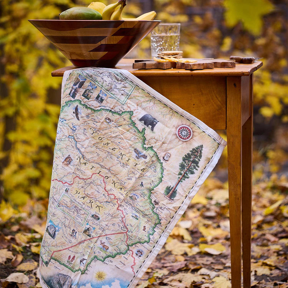 Map-themed table runner on a wooden table with a bowl of fruit and glass in an autumn setting.