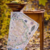 Map-themed table runner on a wooden table with a bowl of fruit and glass in an autumn setting.