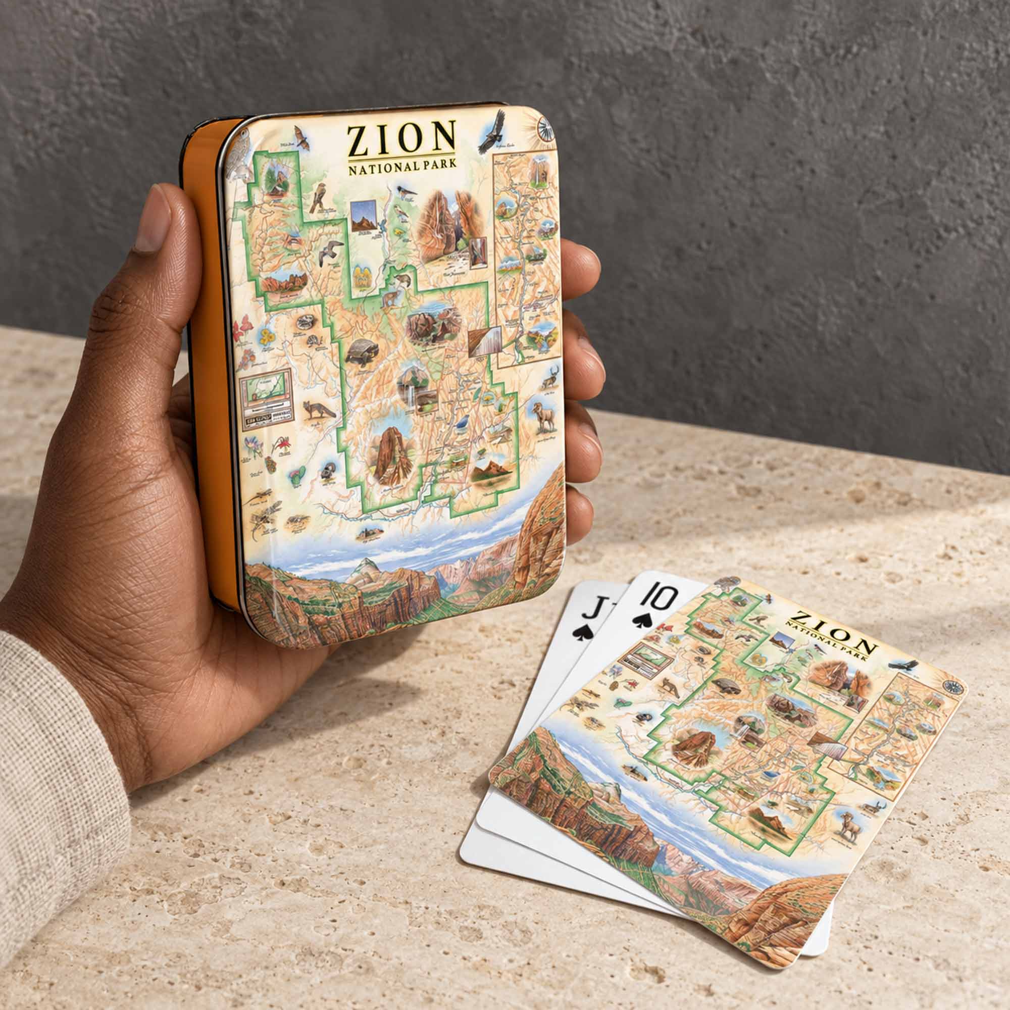 A hand holds a small illustrated metal tin labeled Zion National Park, with matching playing cards placed on a stone surface in front. The product appears to be a Zion National Park themed card deck or set, shown in a real tabletop setting.