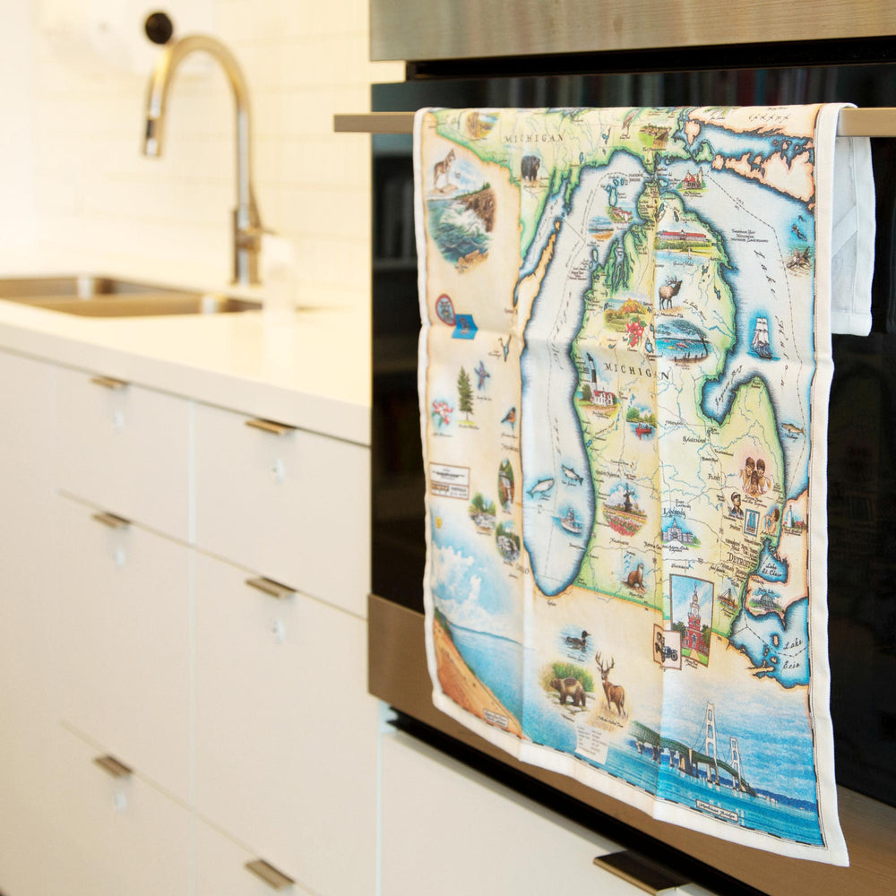 Michigan State Map Kitchen Dishwashing Towel hanging on the kitchen stove handle. The Map features the Great Lakes, Detroit, Ann Arbor, Grand Rapids, and Lansing in earth tones of blue and greens. The tea towel also features  Nature, animals, ducks, deer, fish, moose lighthouses, wolverines, and the Mackinac Bridge.
